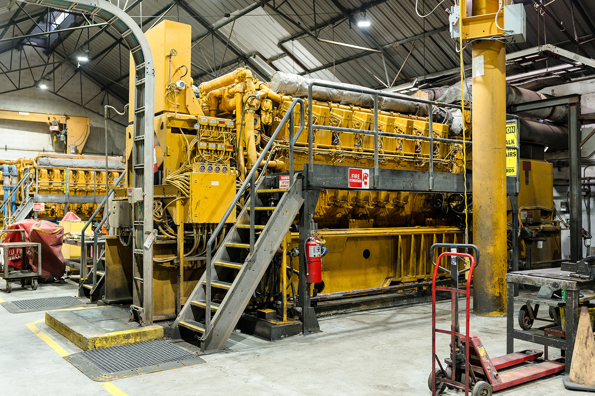 Large industrial generator inside a power plant facility, surrounded by metal stairs, safety equipment, and maintenance tools, with a visible fire extinguisher and safety signs.